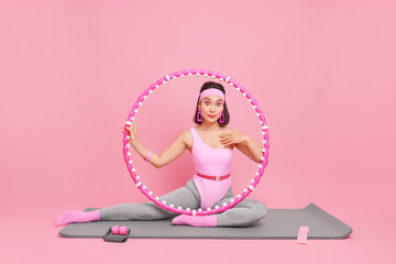 Flexible slim Asian woman poses on fitness mat with hula hoop and other sport equipment does stretching exercises has sports training at home isolated over pink background. Pilates exercising concept