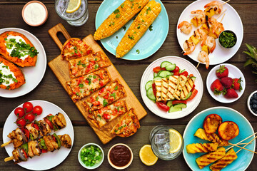 Summer BBQ grill table scene over a dark wood background. Grilled flatbread, chicken and shrimp skewers, stuffed sweet potato, corn, fruit and salad. Top view.