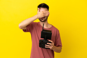 Man holding a drone remote control isolated on yellow background covering eyes by hands. Do not want to see something