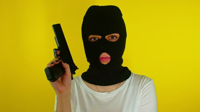 Unrecognizable woman in black balaclava with gun on yellow background. Dangerous criminal in mask with pistol looking at camera.