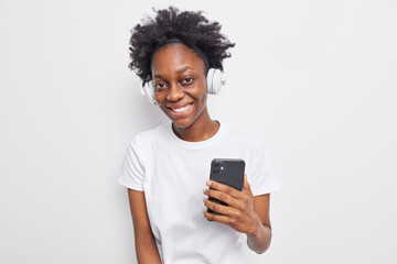 People leisure modern technologies concept. Pretty black curly woman smiles happily holds mobile phone uses application and stereo headphones for listening music dressed in casual white t shirt