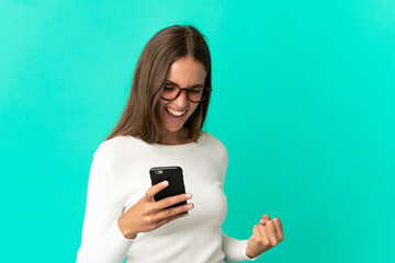 Fototapeta premium Young woman over isolated blue background using mobile phone and doing victory gesture