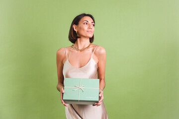Fototapeta premium Woman in silk golden dress on green background holding gift box happy positive excited cheerful posing