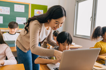 Young adult smiling beautiful Asian teacher helping elementary student boy with laptop in computer classroom. Information Technology class in primary school concept. - 437739444