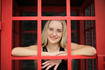 Portrait of Beautiful blonde hair girl on black dress standing in red phone booth against black wall as portrait fashion pose outdoor.