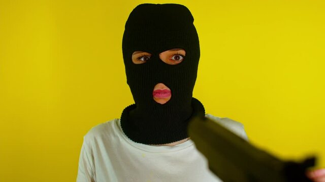 Unrecognizable person points gun at dangerous criminal in black balaclava on yellow background. Unknown scared woman in mask surrenders with her hands up.