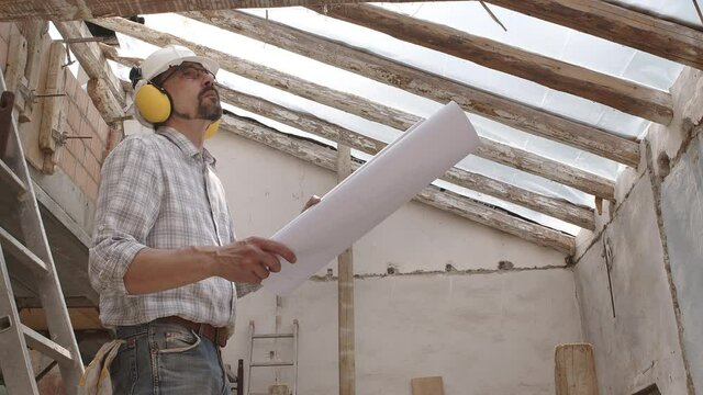 architect man at work, wearing helmet and headphones looking at blueprint , check the construction house project plan, in renovation building site background with the old wooden beams of the roof