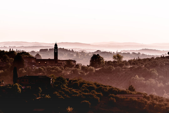 Morning sunrise foggy landscape in Tuscany with silhouette of church bell tower and buildings, near the medieval city of Siena, Italy