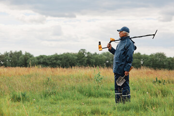 Side view of mature numismatist posing outdoors in field with shovel in hands and metal detector on his shoulder, looking in distance, treasure hunter in meadow.