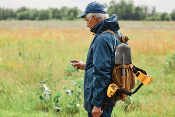 Side view of treasure hunter mature man in jacket and cap holding old coin in hands, looking with interest on his finding, senior numismatist searching with metal detector in field. © sementsova321