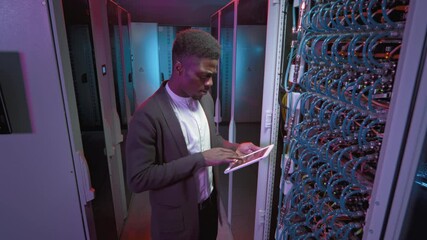 Tracking shot of young African-American male technician with tablet checking servers or other computing equipment in server room of data center