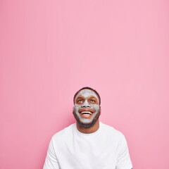 Happy bearded man applies clay mask undergoes beauty treatments focused above feels glad notices something on ceiling dressed in casual white t shirt isolated over pink background blank space