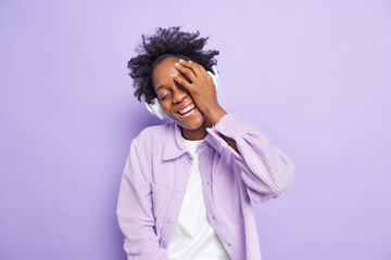 Happy authentic emotions concept. Carefree joyful dark skinned woman with curly hiar makes face palm smiles broadly wears headphones on ears dressed casually isolated over purple background has fun