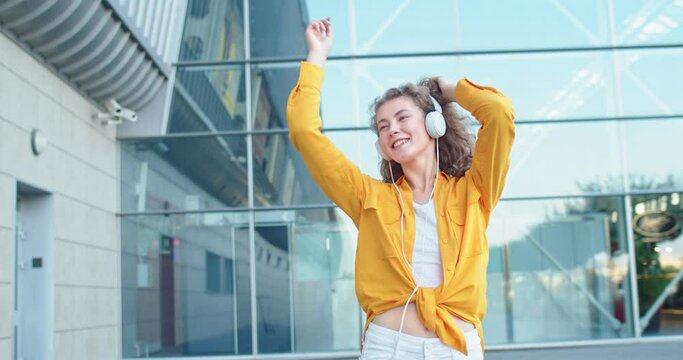 Portrait of Caucasian cheerful young female walking and dancing outdoor listening to fave song outdoor. Happy woman smiling enjoying music on street in city. Leisure, relax, fun concept