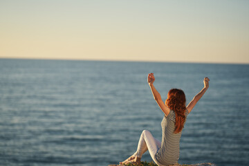 a woman in trousers and a T-shirt sits on the Big Stone near the sea with her hands up