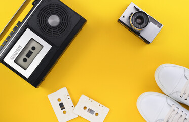 Vintage retro style portable stereo radio with cassette recorder and cassette tapes from around the late 70s with an old camera and white sneakers on a yellow background. Music listening concept. 