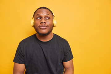 Horizontal shot of thoughtful black man focused above dressed in casual t shirt listens music via headphones thinks about future isolated over yellow background with copy space for your information
