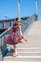 blonde woman in red dress standing outside on stairs and fixing her shoe - obrazy, fototapety, plakaty
