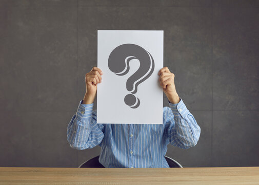Unrecognizable business person holding paper sheet with question mark and asking questionnaire or quiz question. Faceless unknown entrepreneur sitting at desk hiding face behind question mark placard - Powered by Adobe