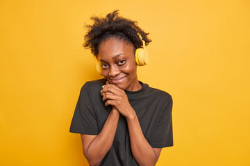 Fototapeta na wymiar Portrait of pleased good looking Afro American teenage girl feels shy keeps hands near face looks satisfied at camera listens music via wireless headphones enjoys free time isolated on yellow wall