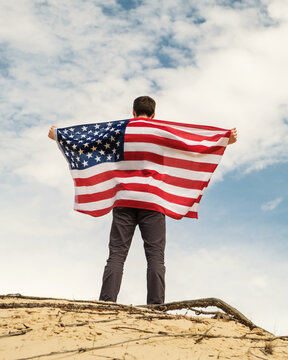 A Man With An American Flag Stands On The Sand, Skies On The Background. Man Is Holding With Both Arms Waving American USA Flag. Fourth Of July Independence Day.