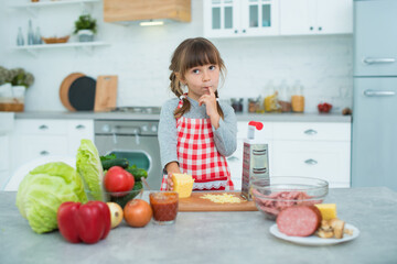 Little cute smiling girl with pigtails and in a checkered cooking apron rubs cheese on a grater, helps to cook pizza in the kitchen
