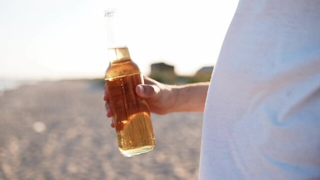 Young handsome caucasian man drinking beer of glass bottle on the beach during sunset in slow motion. Male quenches thirst with lemonade beverage at sandy sea shore. Sun flares, tracking shot.