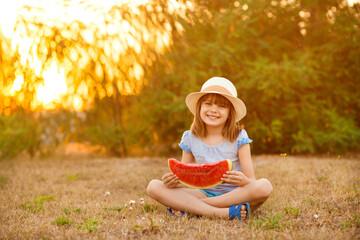 Adorable baby girl in straw hat sit with cross legs on green grass, and eagerly eats juicy...
