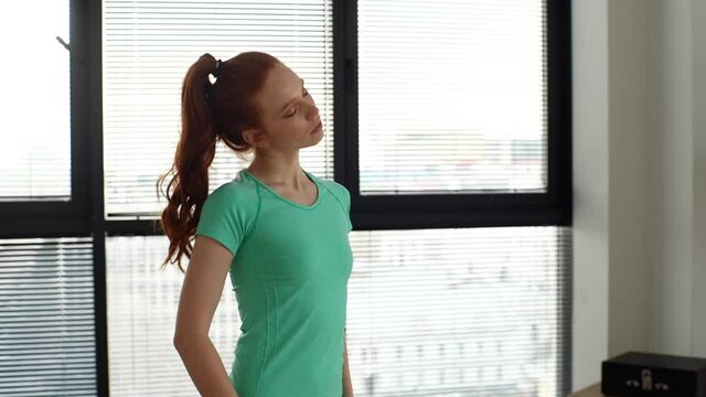 Middle shot portrait of young sporty woman warming up neck before sport fitness workout at home office near window in sunny day. Sporty redhead female warming up before muscles in apartment.