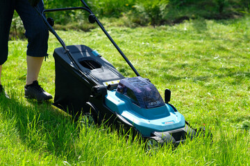 Electric lawnmower cutting green grass. Female gardener with a lawn mower working in backyard