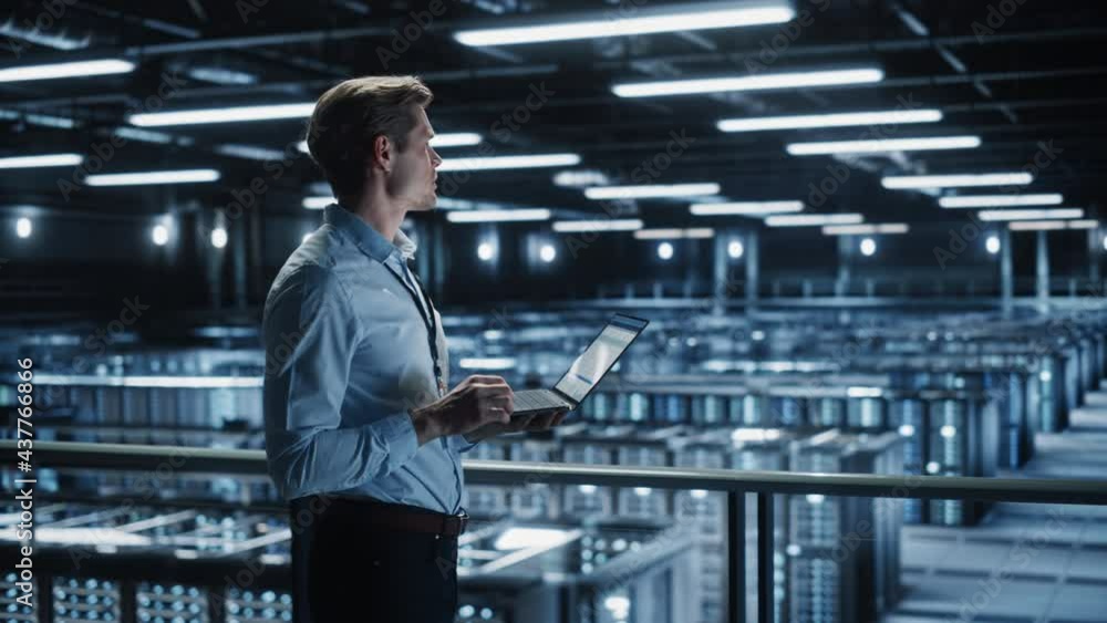 Poster Portrait of IT Specialist Uses Laptop in Data Center. Server Farm Cloud Computing Facility with Male Maintenance Administrator Working. Cyber Security and Network Protection. Medium Tracking Wide
