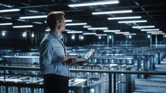 Portrait of IT Specialist Uses Laptop in Data Center. Server Farm Cloud Computing Facility with Male Maintenance Administrator Working. Cyber Security and Network Protection. Medium Tracking Wide