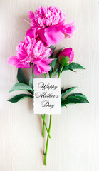 on a light hill wooden background there is a bouquet of three pink peonies with a white rectangular card "Happy Mother's Day". vertical frame