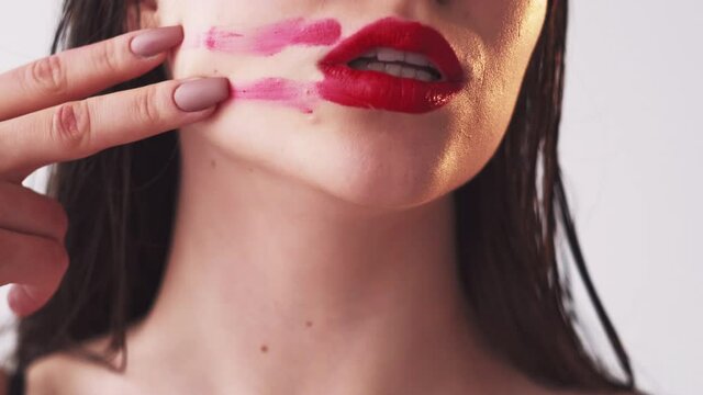 Makeup art. Color cosmetics. Beauty product. Brunette woman smearing smudging red lipstick on face from full lips with two fingers isolated on light.