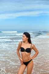 Obraz premium Beautiful luxury slim girl in a black bikini on the beach the ocean. Beauty woman with sexy tanned body, flat stomach, perfect figure. Rest on a tropical island.