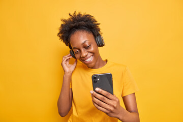 Studio shot of good looking dark skinned woman enjoys favorite playlist listens music via headphones uses smartphone dressed casually isolated over vivid yellow background. Technology concept