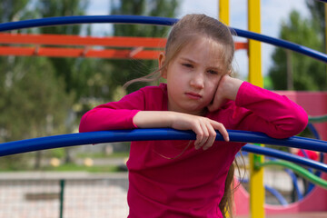 A child girl with blond hair in a pink T-shirt sits offended and sad on the playground. 