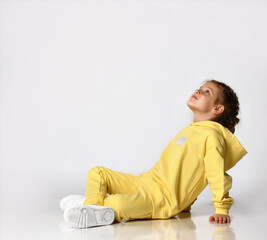 Stylish little girl sitting on a white background and dreamily looking up. Child with a beautiful hairstyle dressed in a yellow sports suit and sneakers looks towards the free space for text.