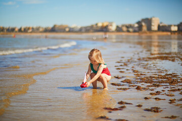 Fototapeta premium Adorable toddler girl in red swimsuit playing with wooden toy boat in water on the beach in Saint-Malo, Brittany, France