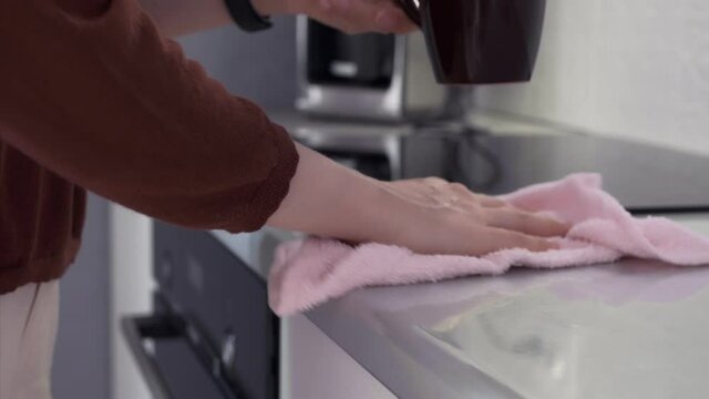 A woman is wiping the enameled light surface of the countertop with a pink kitchen napkin. On the table is a dark mug with white foam on the coffee. The mug is lifted and rubbed underneath.