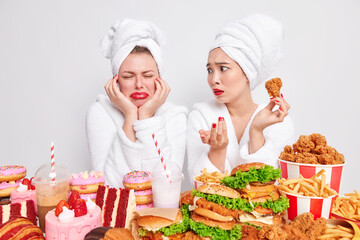 Puzzled Asian woman with red lips looks at her crying friend asks what happened holds fried chicken nuggets dressed in white bathrobe. Two mixed race hungry women afford eating high calorie junk food.