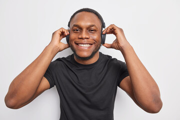 Horizontal shot of handsome hipster guy with dark skin keeps hands on stereo headphones listens favorite track dressed in casual black t shirt isolated over white background. Favotite music.