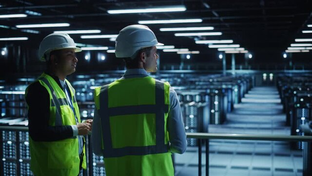 Data Center IT Specialist and System administrator Talk, Use Tablet Computer, Wearing Safety Wests. Server Clod Farm with Two Information Technology Engineers checking Cyber Security. Medium Wide