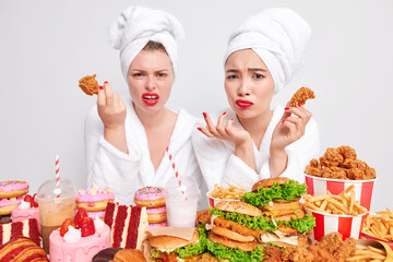 Displeased indignant young women look unhappily at camera hold chicken nuggets eat fast food have bad eating habits surrounded by much cheat meal wear bath towel over head and soft white robe