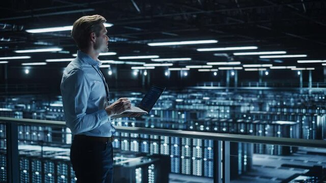 Handsome Smiling IT Specialist Using Laptop Computer in Data Center. Succesful Businessman and e-Business Entrepreneur Overlooking Server Farm Cloud Computing Facility. Medium Wide Shot