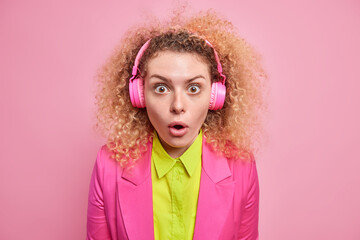 Studio shot of impressed young woman with natural curly hair dressed in formal clothes listens music via headphones cannot believe her eyes isolated over pink background. Human reactions concept