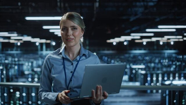 Portrait of Beautiful Smiling IT Specialist Using Laptop Computer in Data Center, Looking at Camera. Succesful Female e-Business Engineer Working in Big Server Farm Cloud Computing Facility. Medium