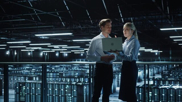 Data Center Female e-Business Enrepreneur and Male IT Specialist talk, Use Laptop. Two Information Technology Engineers work on Bridge Big Cloud Computing Server Farm. Wide Zoom in Two Shot