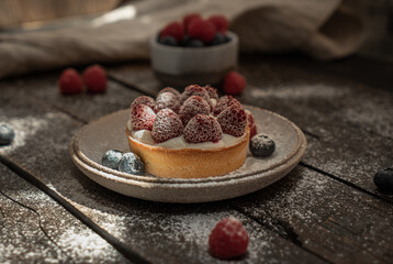 Delicious tartlet with custard, fresh raspberries and sugar powder, served on gray ceramic plate over wooden surface. Summer seasonal sweets. Soft focus.