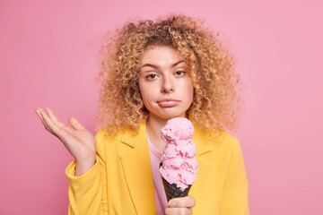 Summer vibes. Hesitant curly haired woman shrugs shoulders with clueless expression eats junk food sweet cold ice cream dressed in yellow jacket isolated over pink background. High calorie dessert